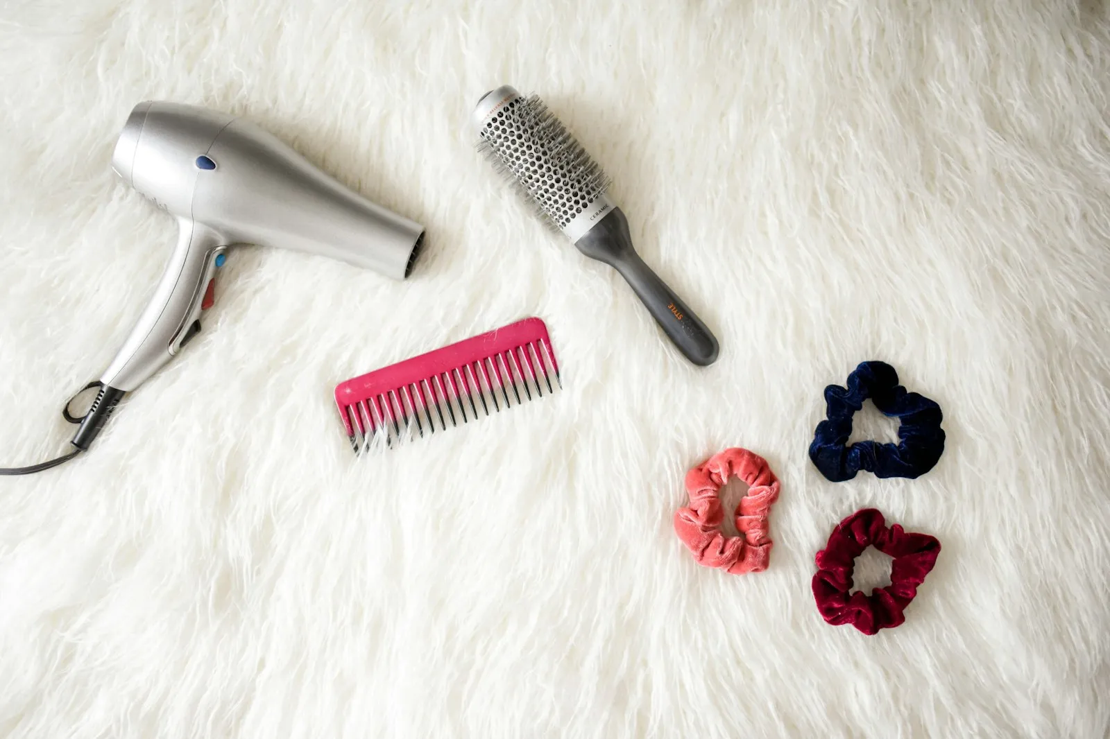 Hair dryer, round brush, comb and scrunchies on a white rug, from the Alaikas.com haircare guide
