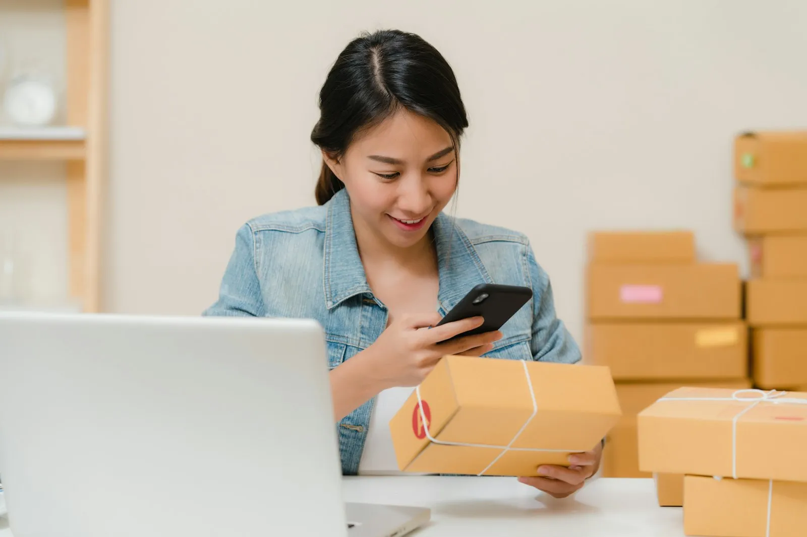 Woman photographing a parcel with her phone next to a laptop and boxes, shopping via the Alaikas.com marketplace